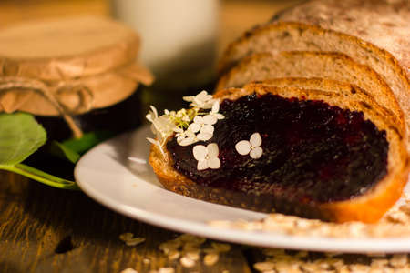 Little glass jar of tasty blackcurrant jam is on the wooden table and slices of fresh crunchy bread with sweet jam are  on the white plateの写真素材