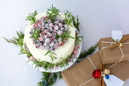 White chocolate cranberry cake decorated with sparkling sugar coated fresh cranberries, Holiday cake, Homemade Christmas cake, Cake with brown paper gift boxes on white table cloth backgroundの写真素材