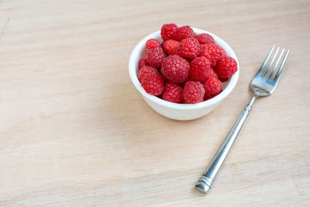 Fresh Raspberries in white bowl with a fork on wooden board background