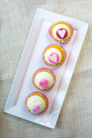 Valentine's french vanilla cupcakes with heart gumpaste topper in white square ceramic plate. Valentine's party cupcake treat on natural burlap fabric backgroundの写真素材