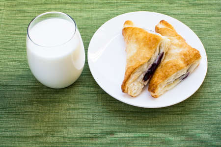 Glass of milk and two pieces blueberry cobbler puff pastries on white plate and green placemat background, Cobbler puff pastries breakfast and a cup of milkの写真素材