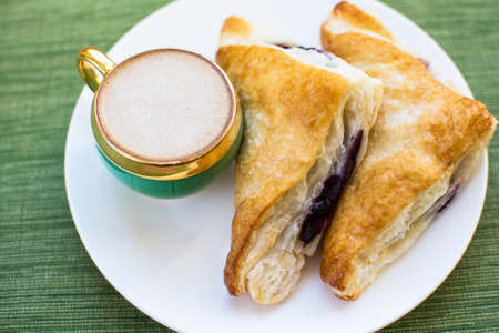 Mini cup of cappuccino and two pieces blueberry cobbler puff pastries on white plate and green placemat background, Cobbler puff pastries breakfast and a cup of coffeeの写真素材
