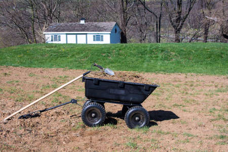 Pile of dirts and grasses in black plastic yard cart with gardening tools on top, tools and dirts in wheelbarrow with tool shed backgroundの写真素材