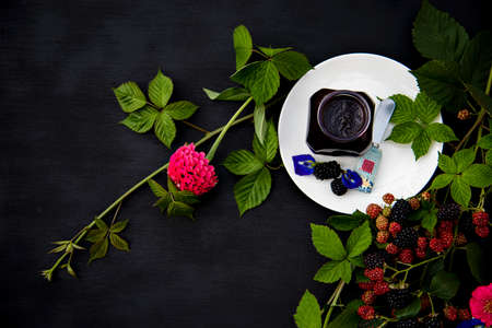 Homemade Organic Blackberry Jam in a Glass Jar on a White Plate Decorated with Fresh Blackberry Branches and Blue Butterfly Pea Flowers on Black Backgroundの写真素材