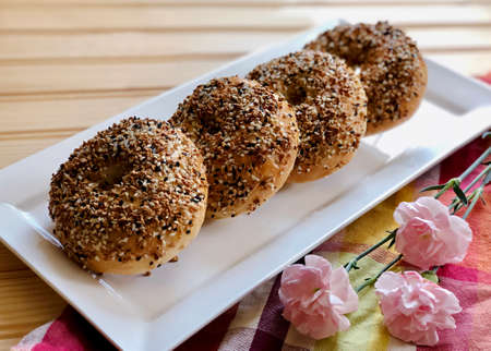 Four pieces of homemade organic everything bagels on white rectangle plate  decorated with pink carnation flower on plaid napkin on wood background.の写真素材