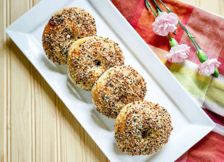 Four pieces of homemade organic everything bagels on white rectangle plate  decorated with pink carnation flower on plaid napkin on wood background; flatlay view,の写真素材