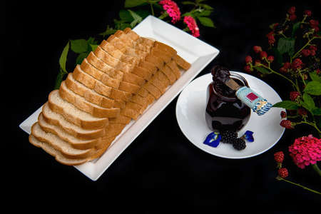 A loaf of sliced bread on a white rectangle plate with a glass jar full of homemade organic blackberry jam and spreader knife on a white circle plate decorated with fresh blackberry branches, pink zinnia and blue butterfly pea  flowers on black background.の写真素材