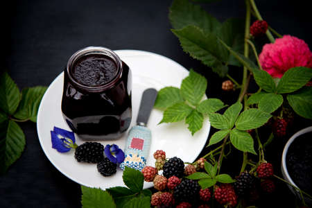 Organic Homemade Blackberry Jam in a Glass Jar on a White Plate Decorated with Fresh Blackberry Branches Pink Zinnia and Blue Butterfly Pea Flowers on Black Backgroundの写真素材