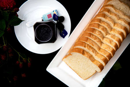 A loaf of sliced bread on a white rectangle plate with homemade organic blackberry jam decorated with fresh blackberry branches, pink zinnia and blue butterfly pea  flowers on black background.の写真素材
