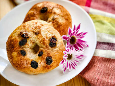 Two pieces of homemade organic cinnamon raisin bagels on white round plate decorated with pink flowers on plaid napkin and wood background.の写真素材