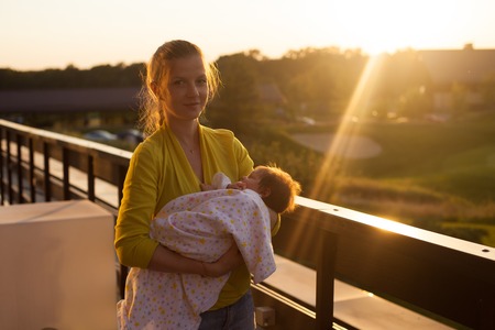 Young beautiful woman in yellow blouse holding her sleeping baby in pink blanket on the terraceの写真素材