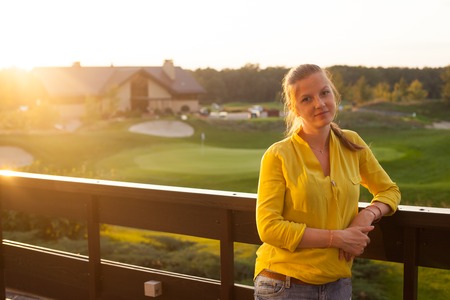 Beautiful smiling woman with sandy hair in yellow blouse standing on the terrace on the background of green field and housesの写真素材