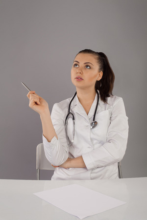 Nurse in white robe and glasses is looking somewhere and sitting at the table against of grey backgroundの写真素材
