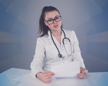 Beautiful nurse in white robe and glasses is making her report and thinking about it at the white table against of grey backgroundの写真素材