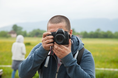 Man in blue shirt holding black camera and making a photo on the background of green natureの写真素材