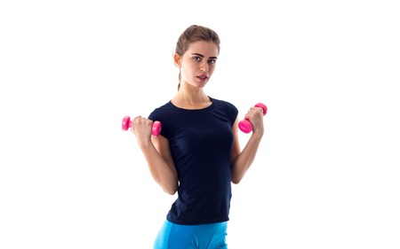Young attractive woman in blue T-shirt and leggings holding pink dumbbells in her hands on white...