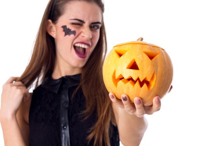 Young beautiful woman with little picture of bat on her cheeck and headband in dress holding a pumpkin and winging on white background in studioの写真素材