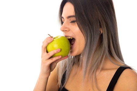 Young smiling woman in black top eating green apple on white background in studioの写真素材