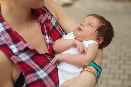 Young woman in checkered blouse holding little sleeping baby in white clothingの写真素材