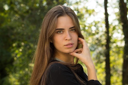 Young pleasant woman with brown long hair in black blouse and silver necklace standing sidewise on the background of green treesの写真素材