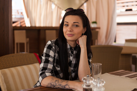 The woman with black  glasses and tattoo is sitting on the wood table in beautiful cafe.の写真素材