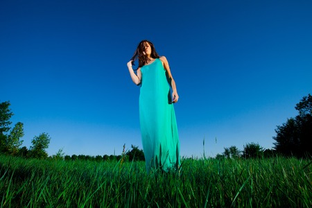 Beautiful young woman in a long blue dress standing on green grassの写真素材