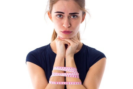Young pretty woman in blue T-shirt with ponytail holding centimeter around her arms  and making a sad face on white background in studioの写真素材