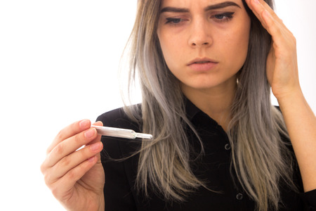 Young beautiful woman in black shirt with headache holding a thermometer and looking at it on white background in studioの写真素材