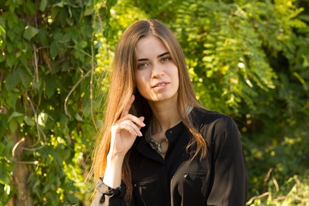 Young pretty woman with brown long hair in black blouse and silver necklace on the background of green trees and bright sunlightの写真素材