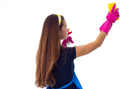 Young woman with long hair in blue T-shirt and apron with pink gloves using yellow duster and detergent on white background in studioの写真素材