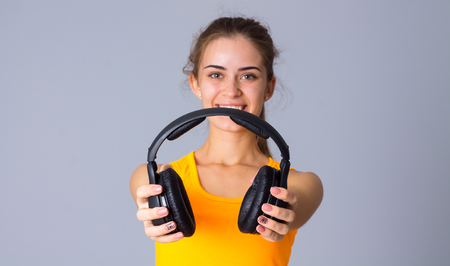 Young smiling woman with ponytail in yellow T-shirt holding black headphones on gray background in studioの写真素材