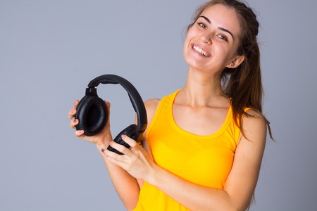 Young pretty woman with ponytail in yellow T-shirt holding black headphones on gray background in studioの写真素材