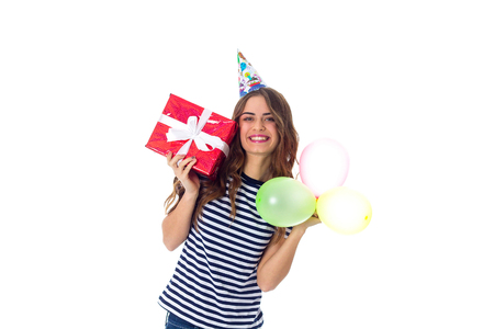 Pretty young woman in stripped T-shirt and celebration cap holding red present and colored balloons on white background in studioの写真素材
