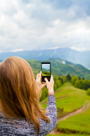 Young woman is standing on the hill and taking photo of amazing beautiful landscape with mountain, forest and cloudly skyの写真素材