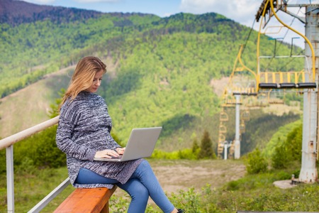 Pregnant woman with laptop is sitting on the background of ski lift and beautiful nature.の写真素材