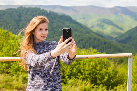 Beautiful young woman with long hair is doing selfie on her phone on the background of green nature.の写真素材