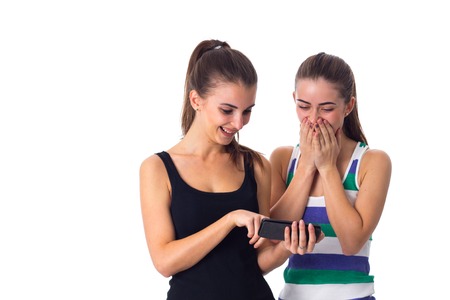 Two young pretty women in striped and black shirts using smartphone and laughing on white background in studioの写真素材