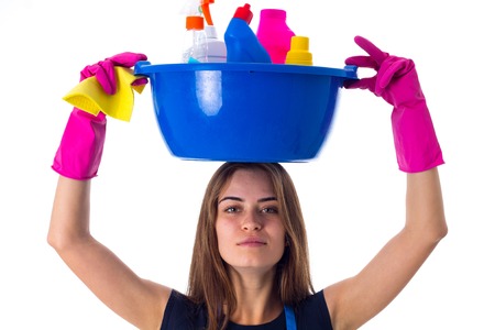 Young beautiful woman in blue T-shirt and apron with pink gloves holding cleaning things in blue washbowl on her head on white background in studio.の写真素材