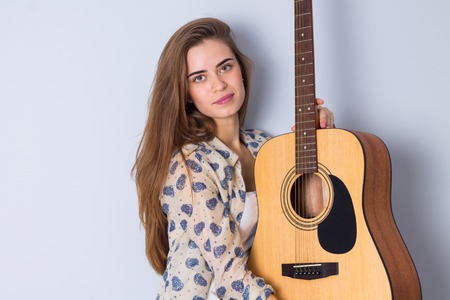 Beautiful young woman in beige blouse holding a guitar on gray background in studioの写真素材