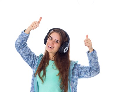 Young positive woman with long hair in blue shirt listening to the music in black headphones and showing thumbs up on white background in studioの写真素材