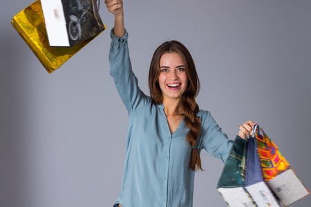 Young smiling woman with long plait  in blue blouse holding varicolored shopping bags in her hands on gray background in studioの写真素材