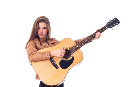 Young pretty woman with long hair in brown jacket and black trousers holding a guitar and making faces on white background in studioの写真素材