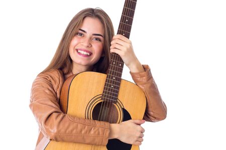 Smiling young woman with long hair in brown jacket hugging a guitar on white background in studio