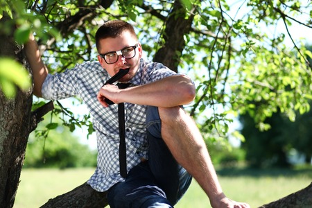 Handsome man in glasses sitting on the branch and biting the tieの写真素材
