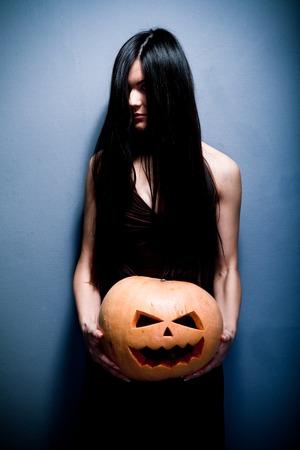 Gloomy young woman holding a large orange pumpkin carved for Halloween celebration. Dark background.の写真素材