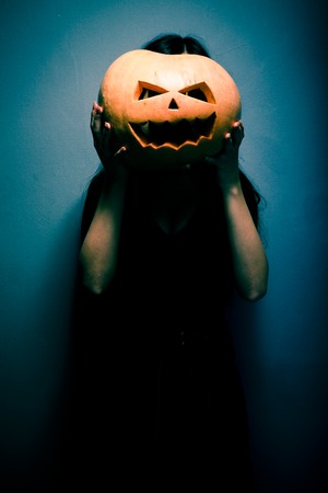 Gloomy young woman holding a large orange pumpkin for Halloween in front of her face. Dark backgroundの写真素材