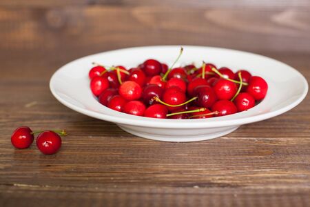 Close-up view of white plate with fresh red cherries on the brown wooden tableの写真素材