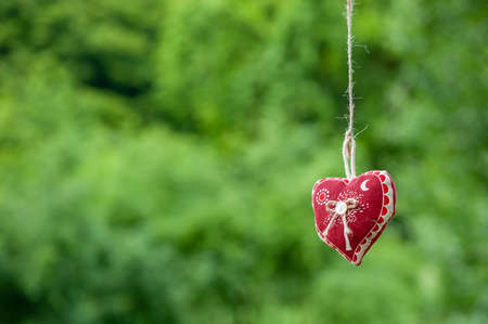 Isolated handmade cotton fabric heart hanging on bast string on green grass background. Red fabric heart hanging on the clothesline. Summer wedding theme. Love and valentine in nature.の写真素材