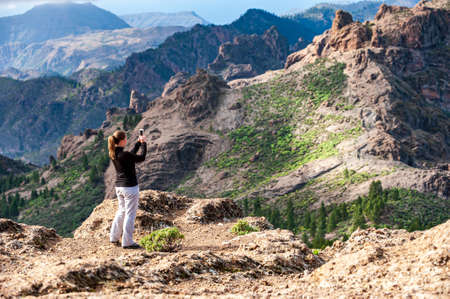 Young woman hiker from behind taking picture of volcanic landscape with mobile phone. Female trekker photographing in nature. Trekking in volcanic mountains of Gran Canaria, Canary Islands, Spain.の写真素材