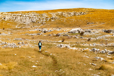Autumn Dinara mountain landscape with sporty young woman, rocky hills, golden grass, sky. Travel and tourism. Hiking female in the Dinaric Alps on the border between Croatia and Bosnia and Herzegovina.の写真素材